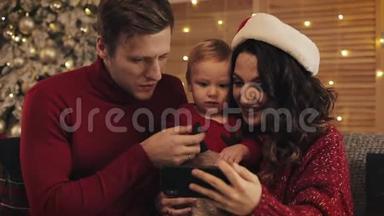 <strong>爸爸妈妈</strong>和小男孩的年轻快乐家庭在圣诞树附近的沙发上坐着，背景下有明亮的灯光